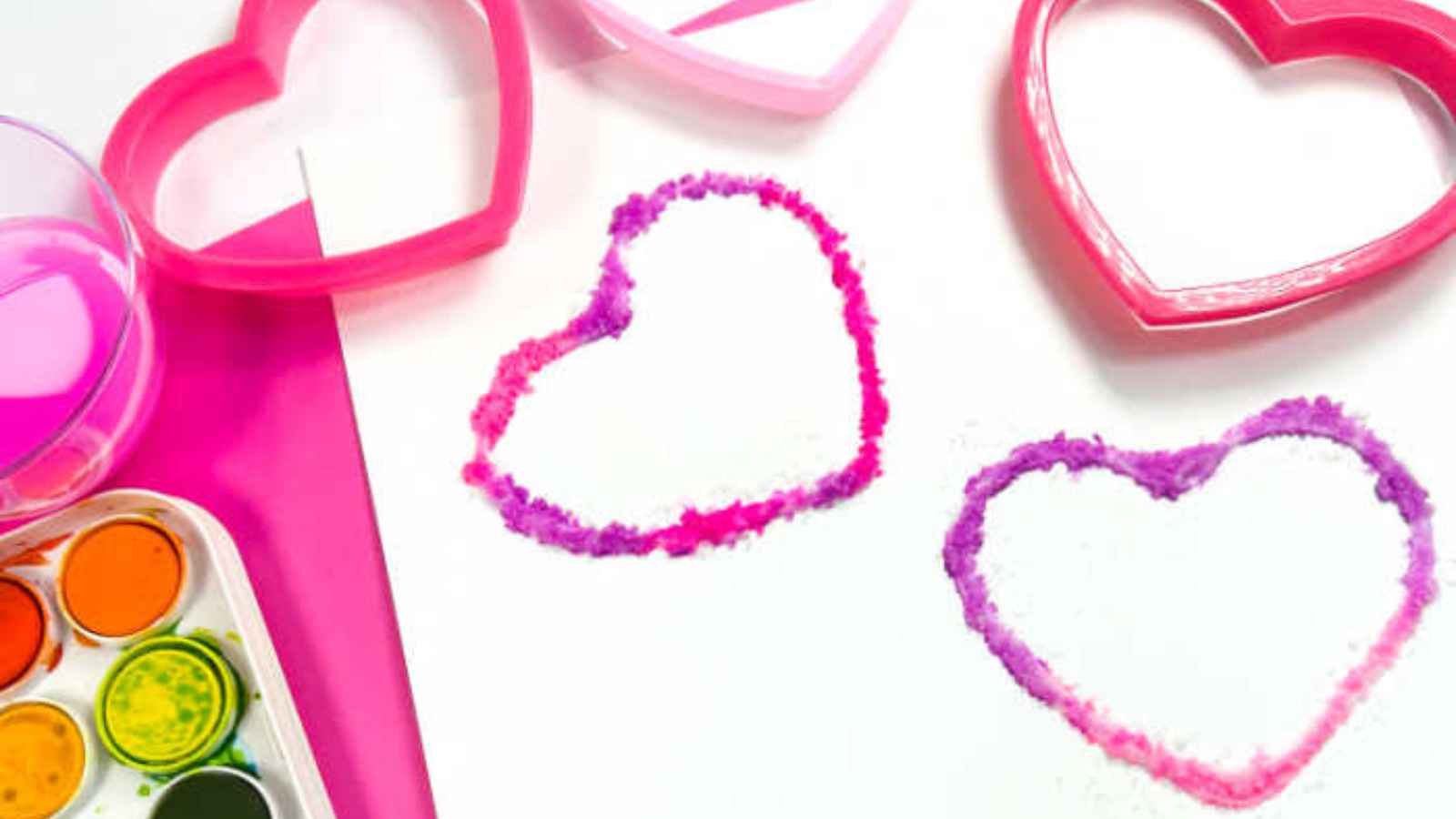 Two heart shapes outlined in pink and purple on paper, with heart-shaped cookie cutters and watercolor paints nearby.