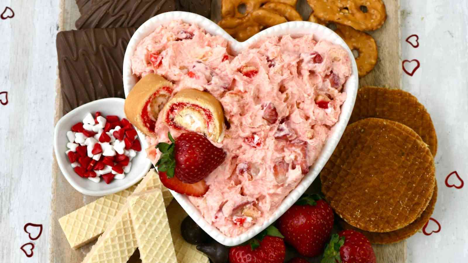 Heart-shaped bowl with strawberry dip surrounded by wafers, chocolate, pretzels, strawberries, and a small bowl of candy hearts on a wooden board.