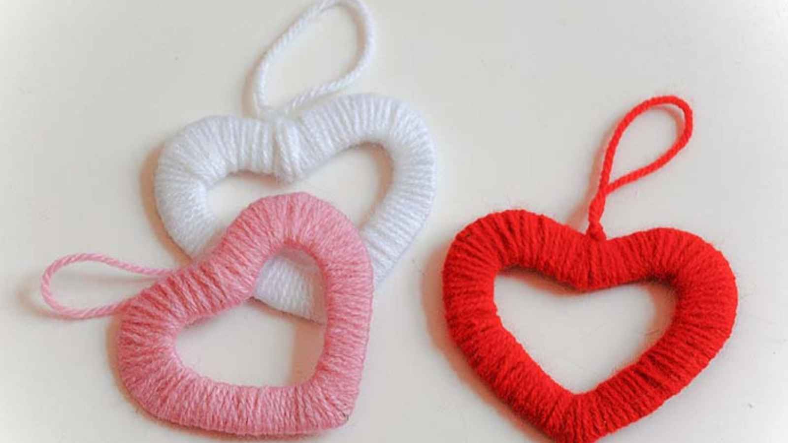 Three yarn-wrapped heart shapes in white, pink, and red, each with a loop at the top, displayed on a light surface.