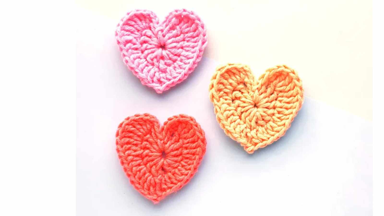 Three crocheted heart shapes in pink, orange, and red are arranged on a light background.