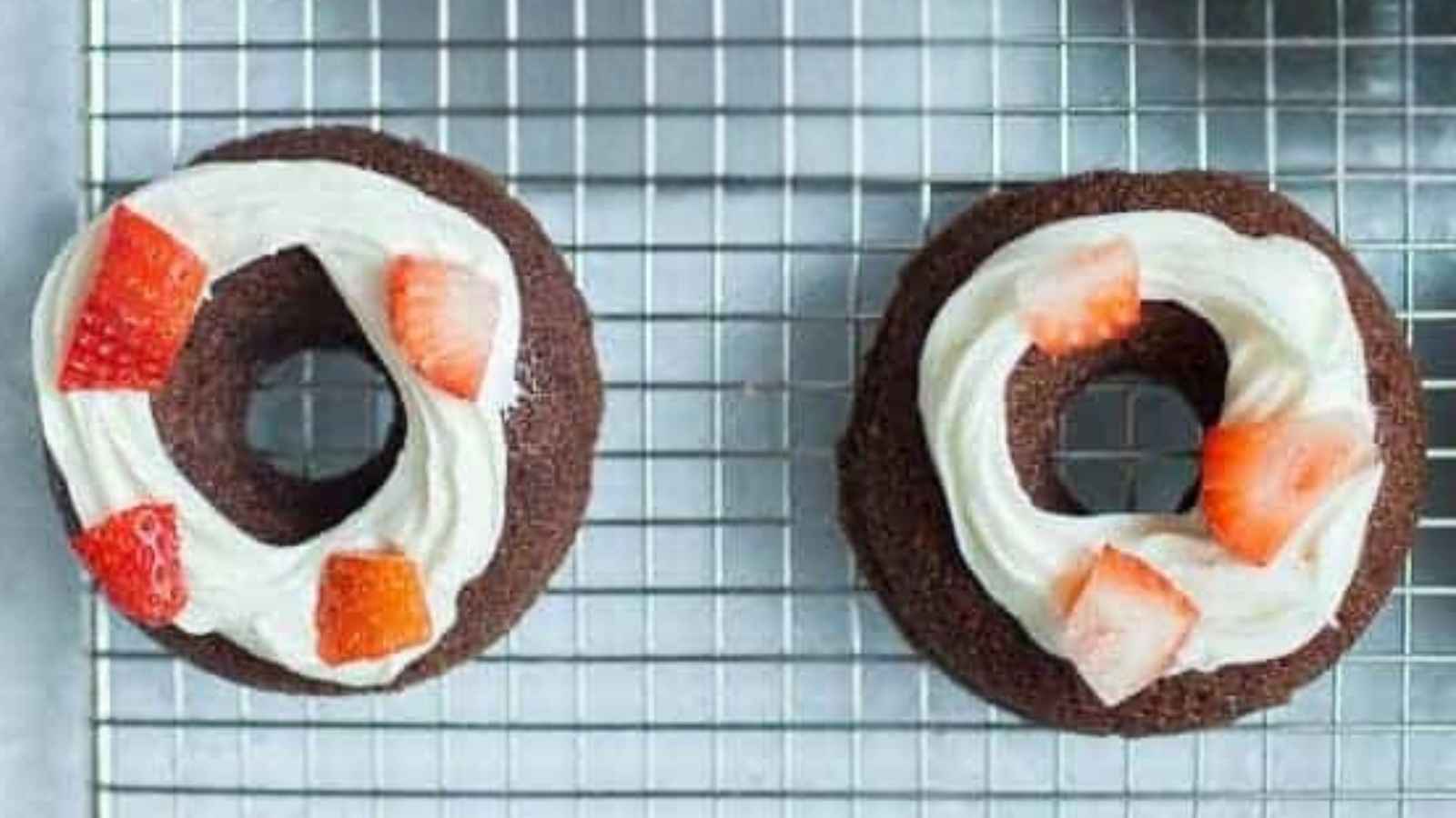 A cooling rack with four chocolate brownie donuts topped with white frosting and small pieces of fresh strawberries. A red and white striped cloth is partially visible in the background, along with a piping bag with a blue tip on the side.