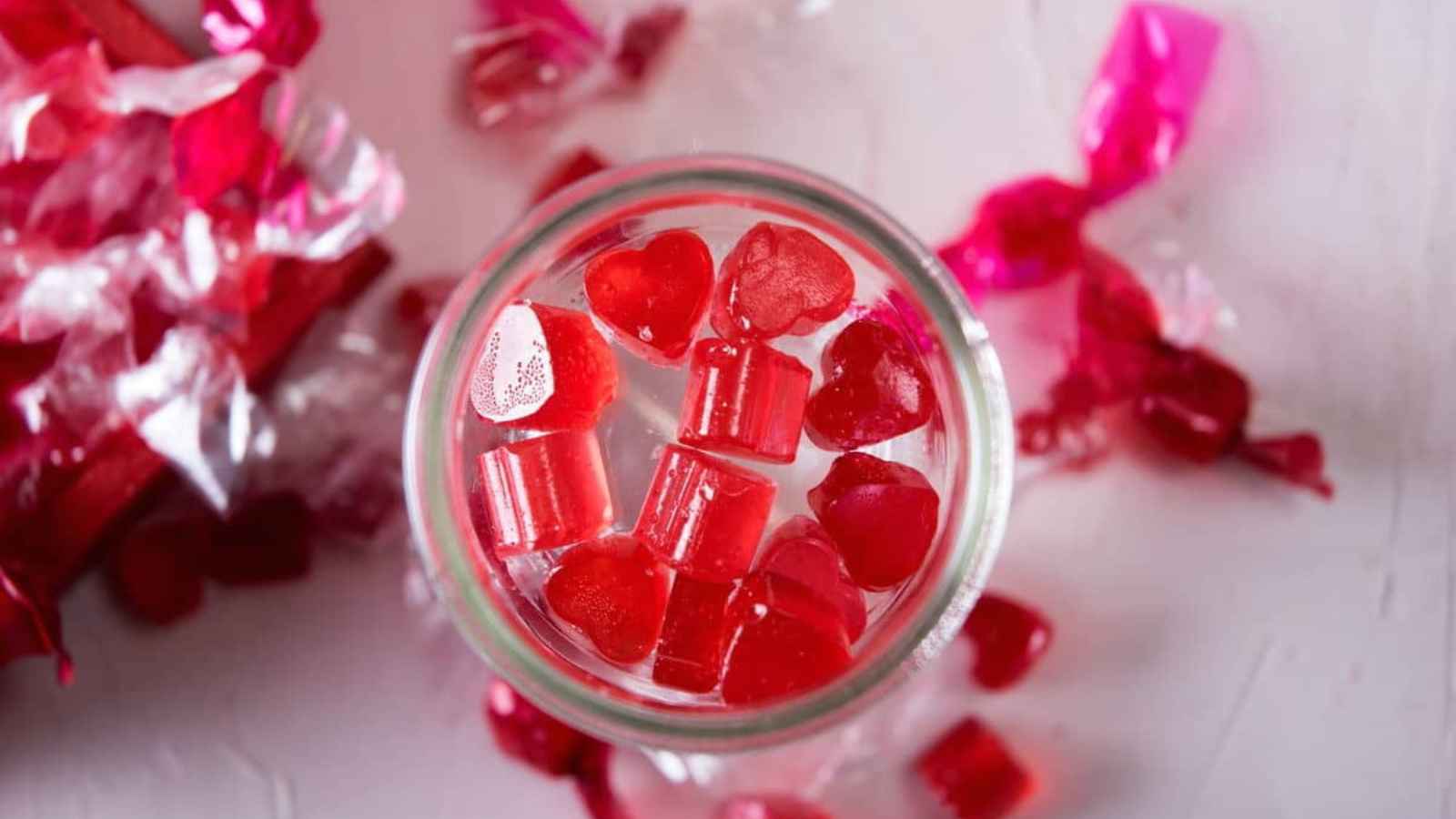 A jar filled with red heart-shaped candies, surrounded by opened pink wrappers on a white surface.