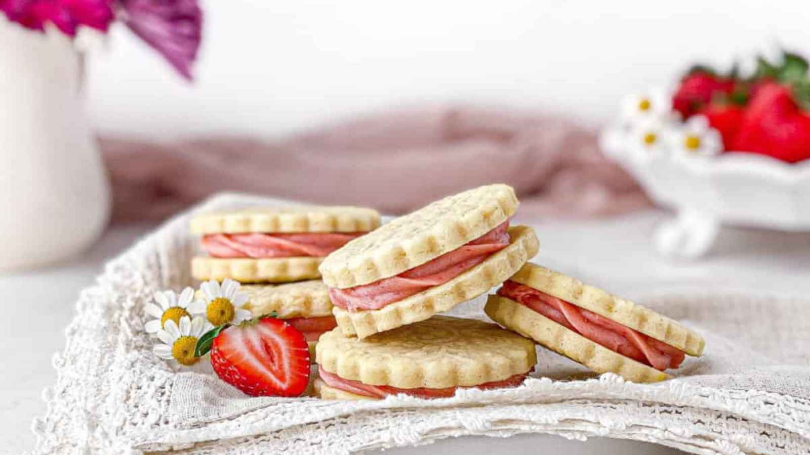 Cookies filled with pink cream are stacked on a lace cloth. Two daisies and a sliced strawberry are in the foreground, with strawberries in a bowl in the background.