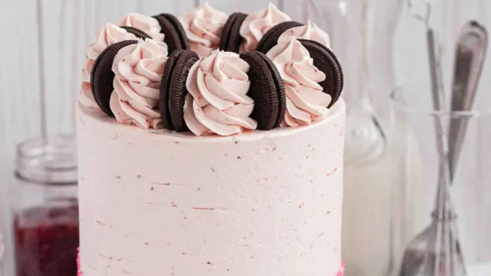 A tall cake with white frosting, topped with swirls of pink cream and chocolate sandwich cookies.