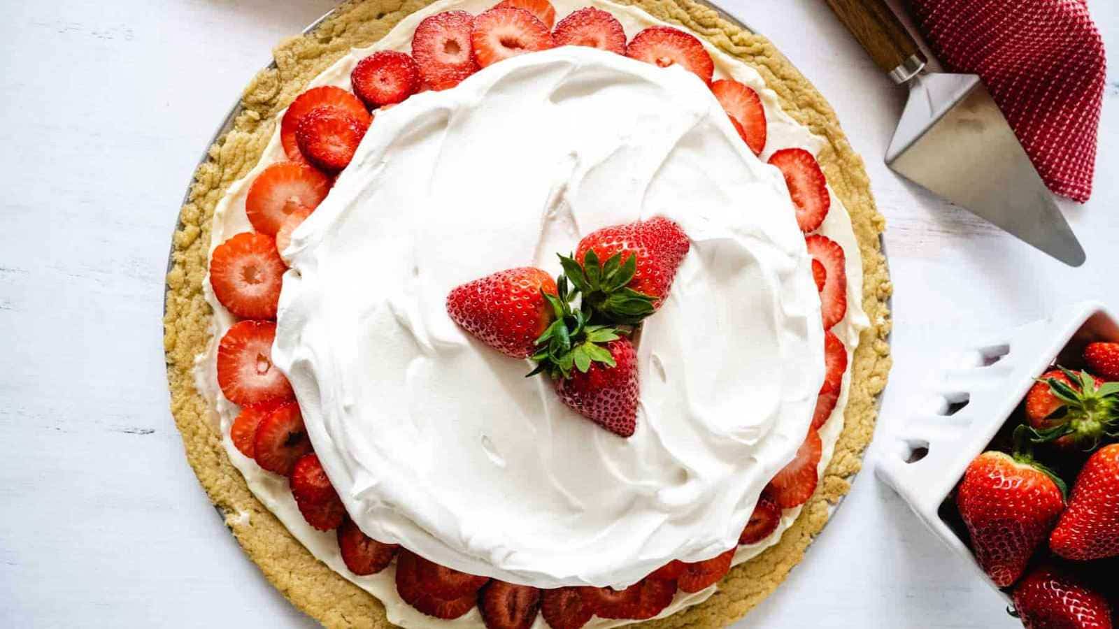 A strawberry dessert pizza topped with whipped cream and strawberries, surrounded by a pizza cutter and a basket of strawberries on a white surface.
