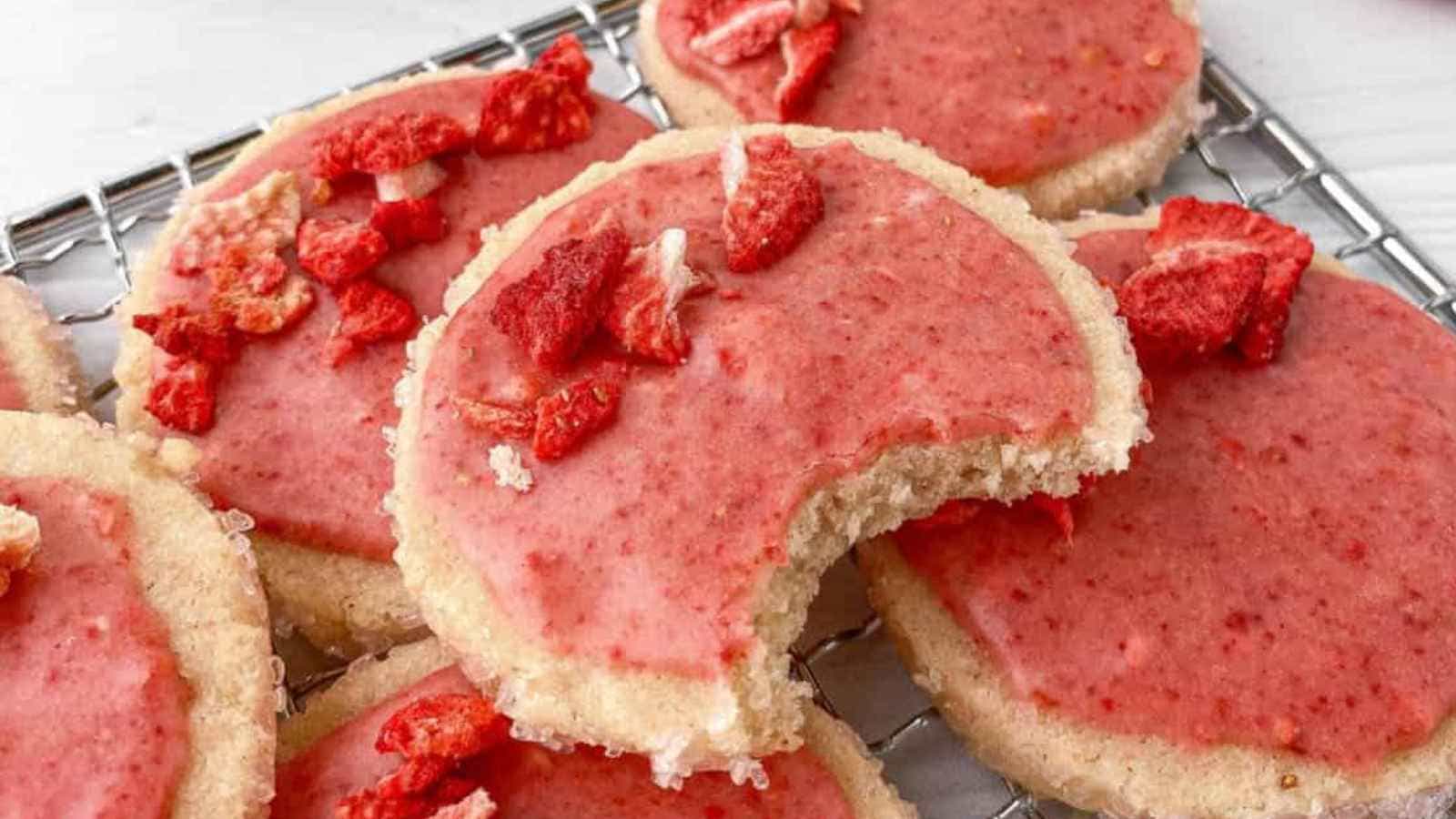 Cookies with pink frosting and dried strawberry pieces on a cooling rack. One cookie has a bite taken out of it.