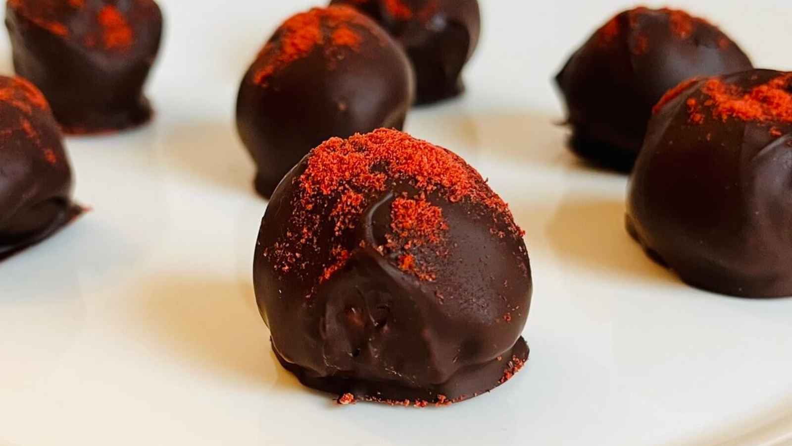 Close-up of dark chocolate truffles topped with red powder, arranged on a white plate.