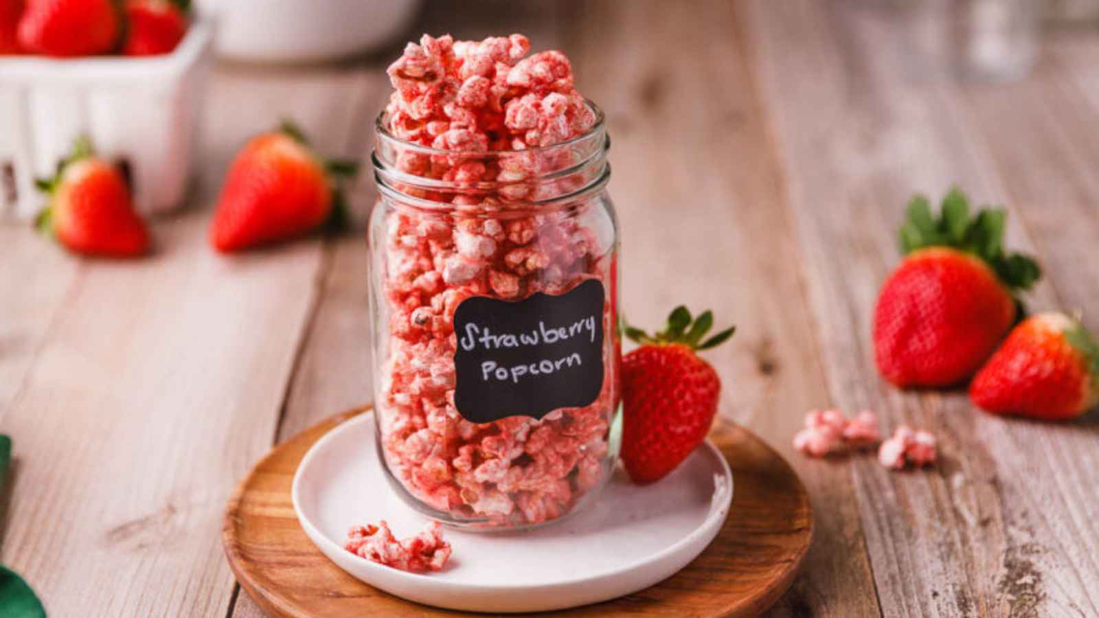 A jar of strawberry-flavored popcorn on a plate, surrounded by fresh strawberries on a wooden table.