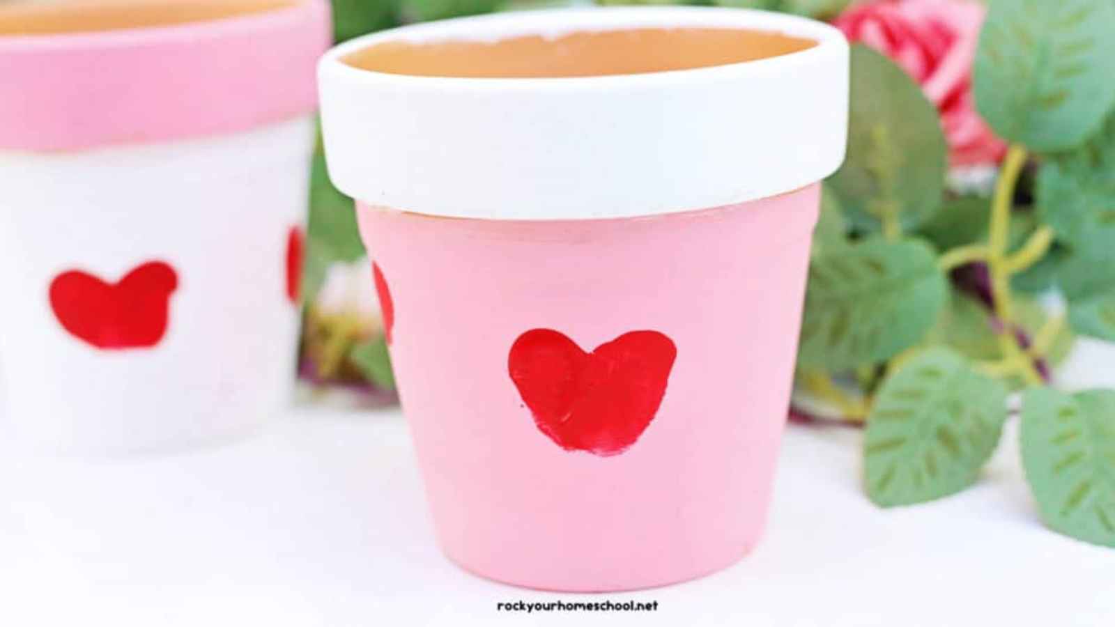 A pink and white flower pot with a red heart design on it, surrounded by green leaves.