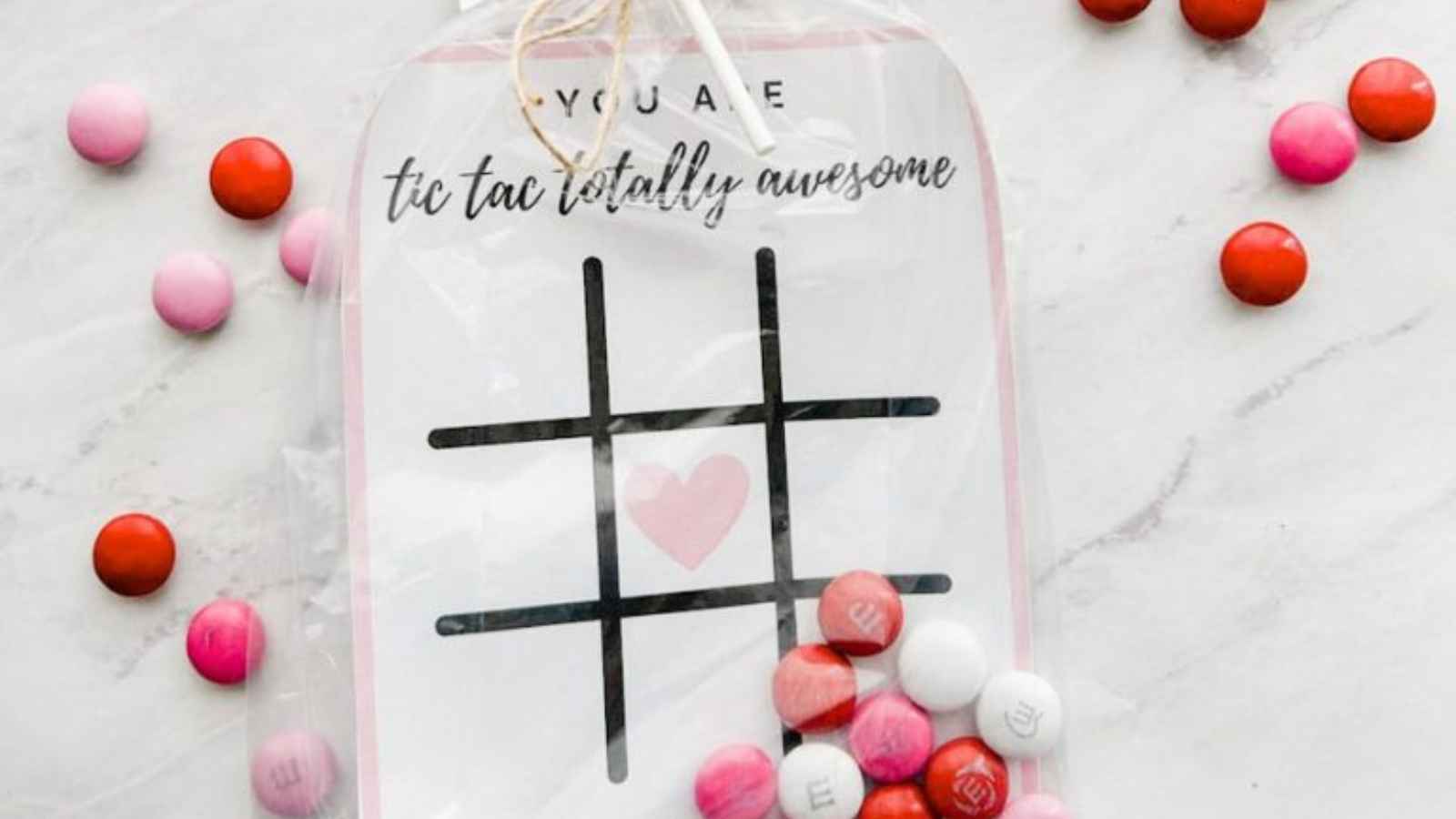A tic-tac-toe board with a heart in the center is surrounded by red, pink, and white candies on a marble surface.