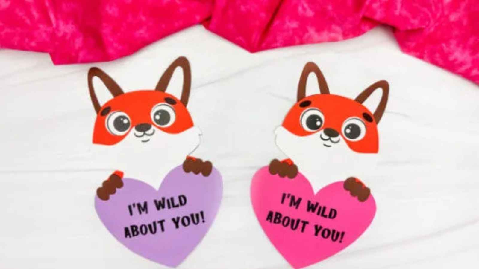 Two cartoon foxes holding purple and pink heart-shaped signs that read, "I'm wild about you!" on a white surface with a pink fabric background.