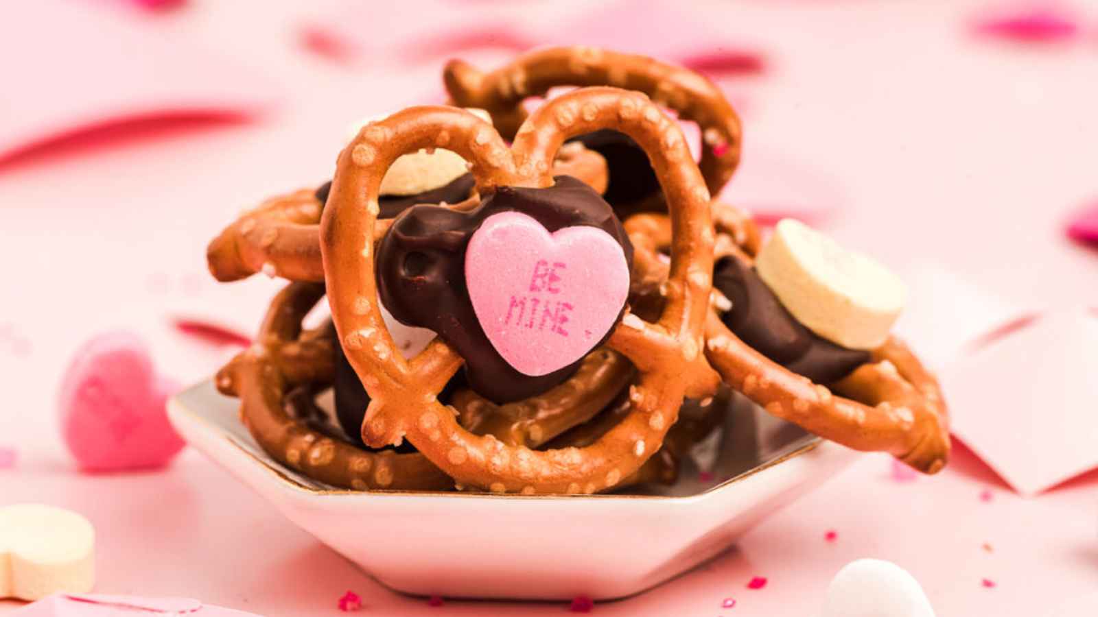 A small dish filled with heart-shaped pretzels, chocolate, and candy hearts, one reading