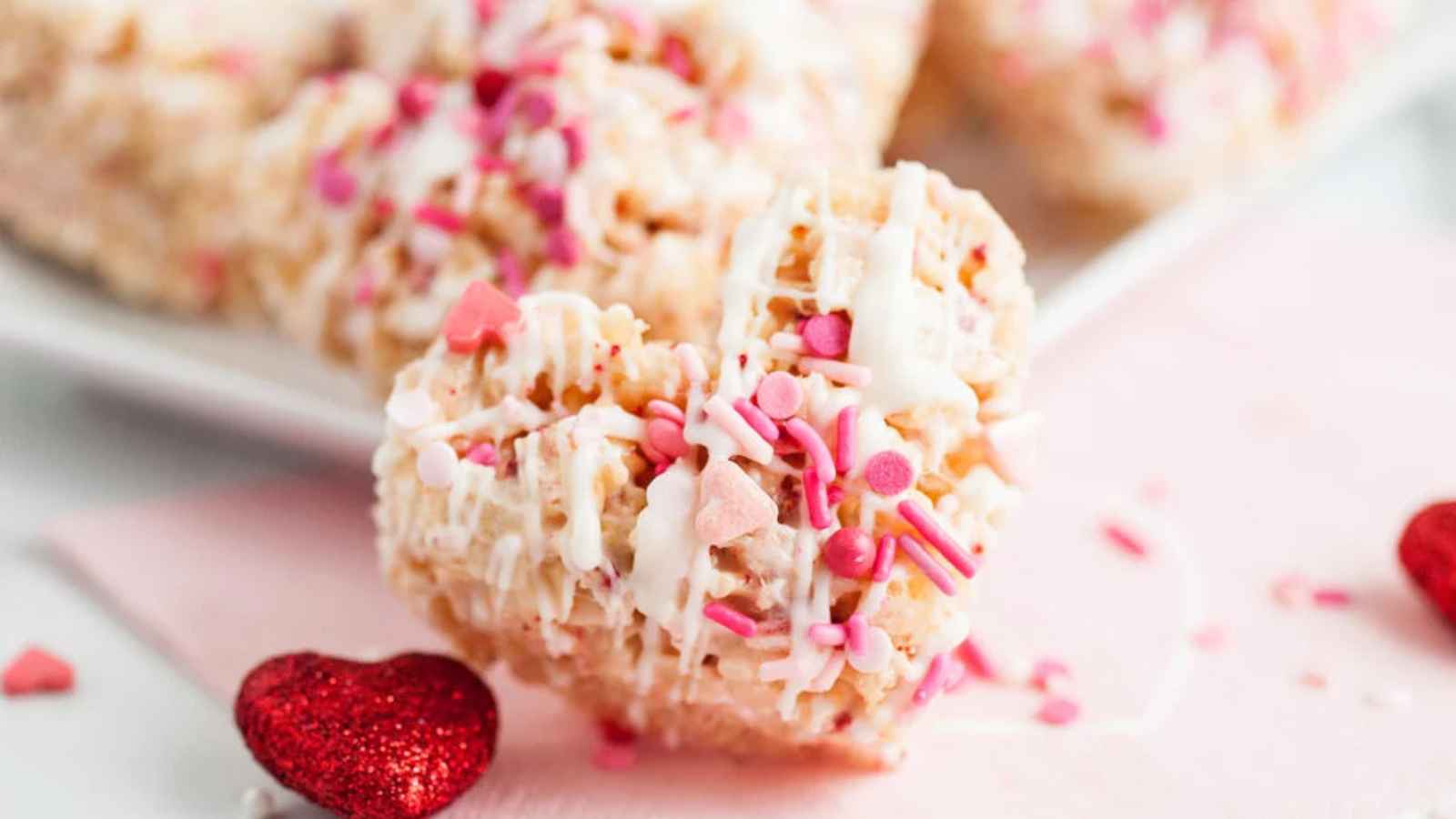 Heart-shaped dessert with pink and white icing, topped with sprinkles, on a pink surface.