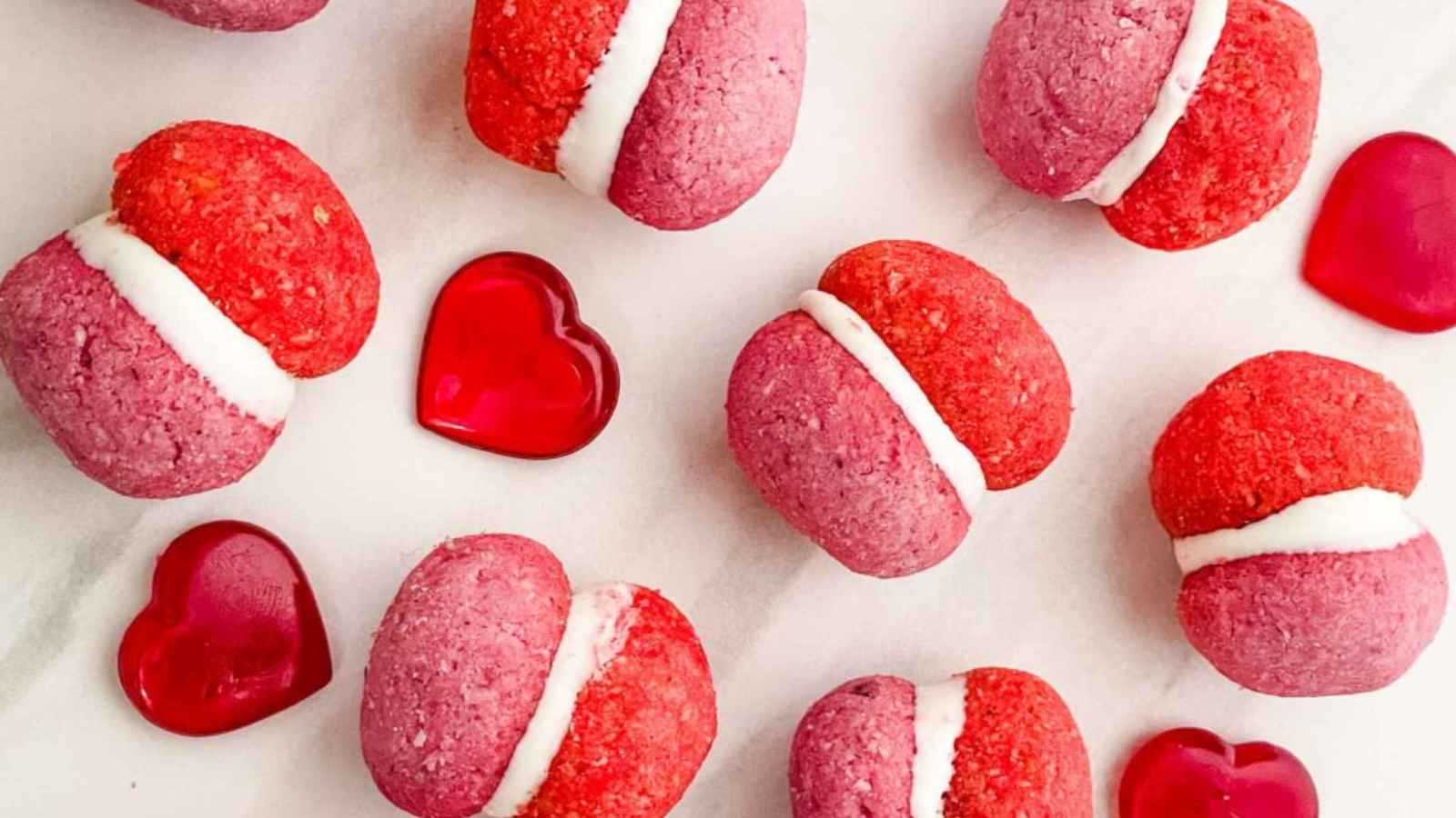 Pink and red Valentine’s Day Baci di Dama Cookies with white filling, surrounded by red heart-shaped jelly candies on a white surface.