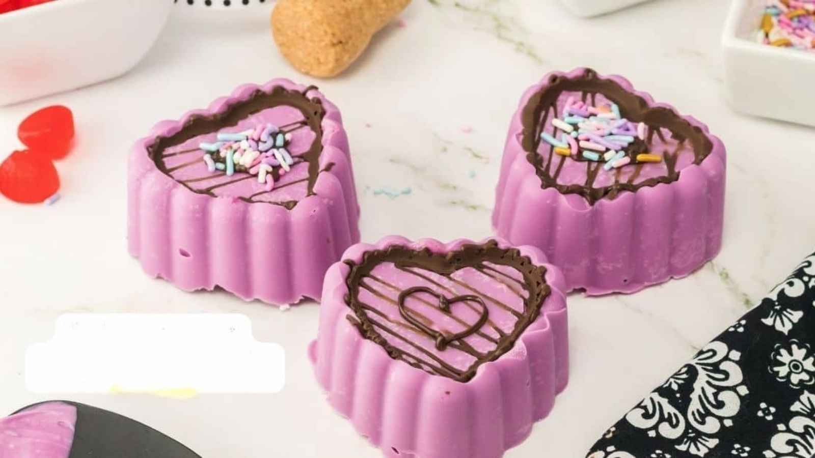Three heart-shaped purple cakes with chocolate icing and colorful sprinkles on top, placed on a white surface.