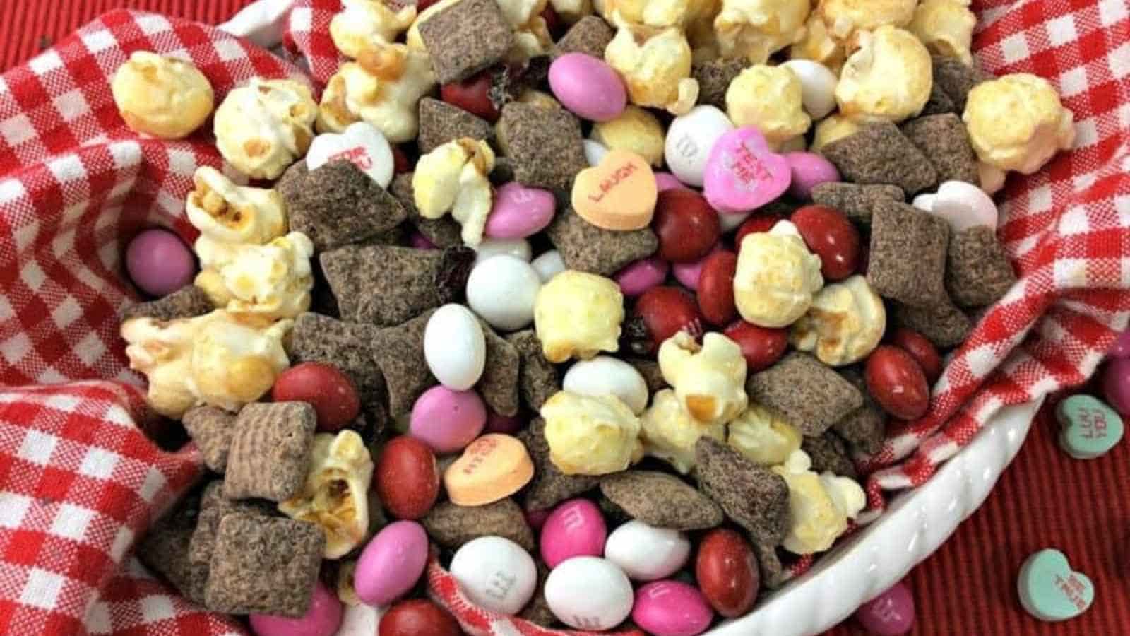 A bowl contains a mix of popcorn, chocolate cereal squares, candy-coated chocolates, and heart-shaped candies on a red and white checkered cloth.