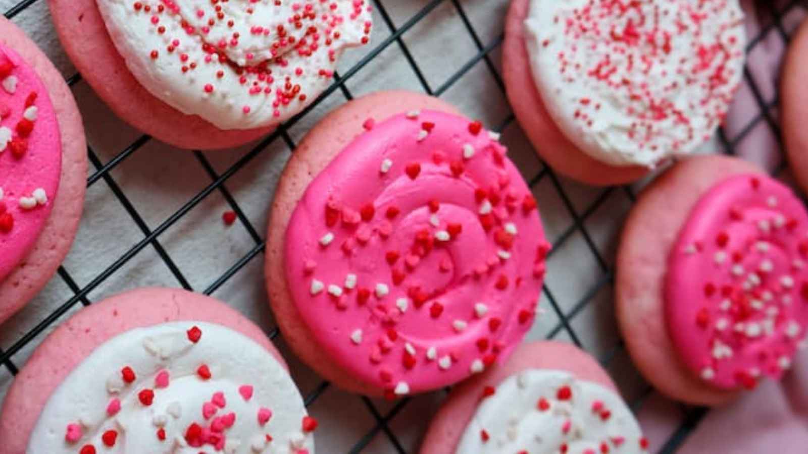 Pink and white frosted cookies with sprinkles on a cooling rack.