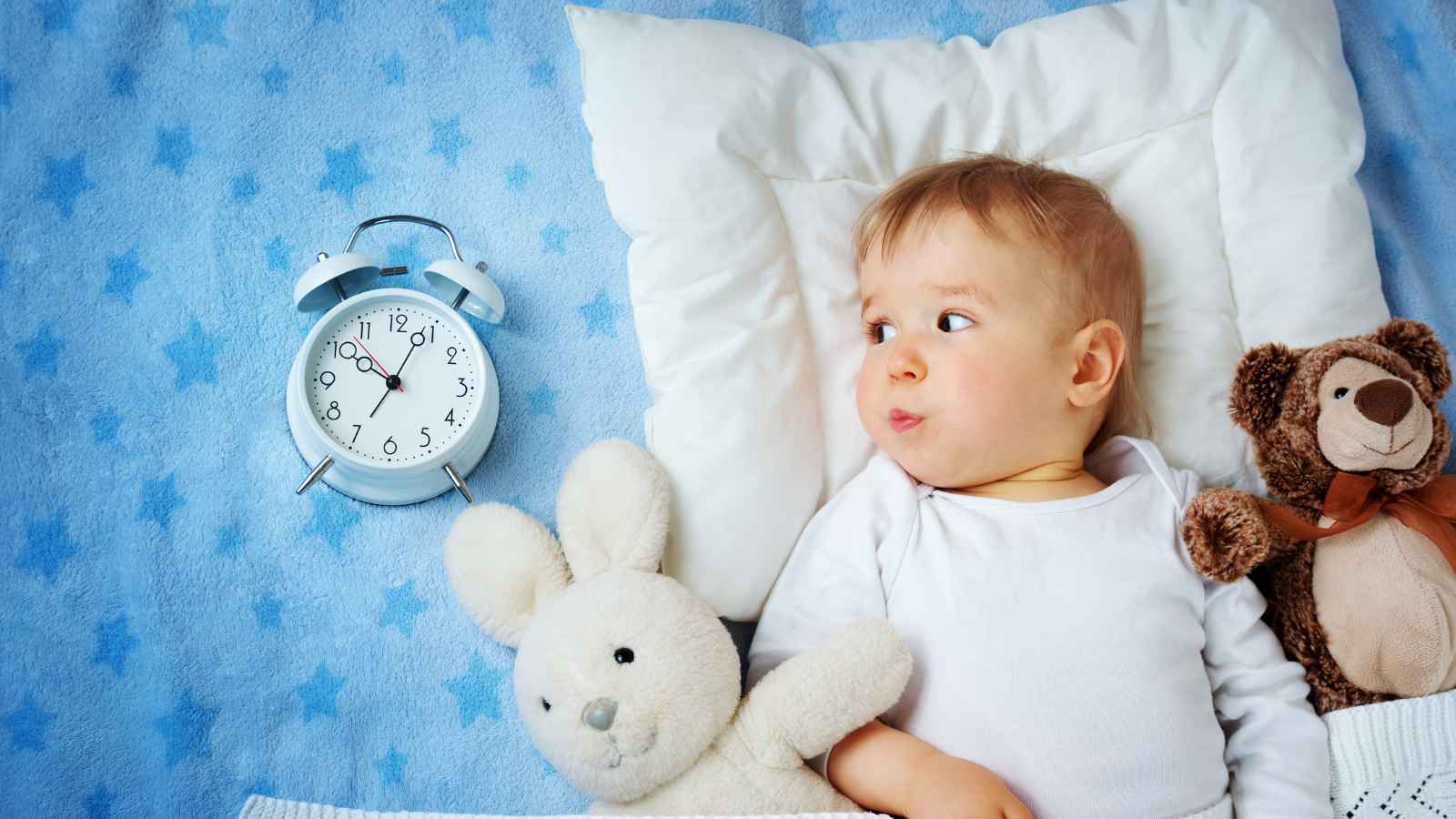 A baby rests on a blue star-patterned blanket, surrounded by a white stuffed rabbit, a brown teddy bear, and an alarm clock.