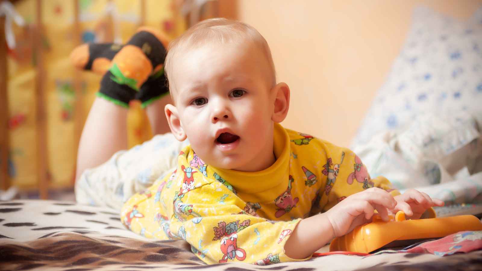 A toddler in a yellow shirt with colorful patterns lies on a bed, clutching a toy and gazing ahead. Features a cozy crib and scattered pillows in the background.