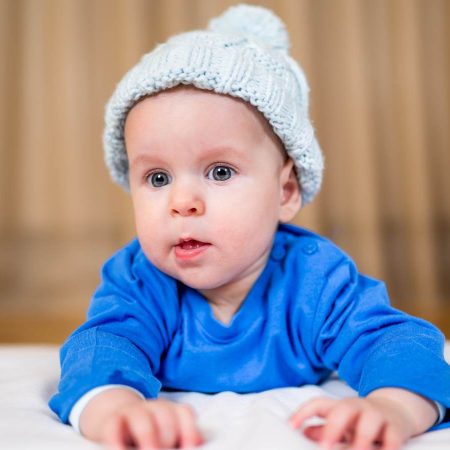 A baby in a blue outfit and knit hat lies on a white blanket, looking forward.