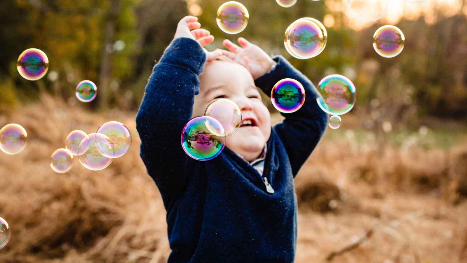 A young child in a navy sweater, joyfully reaches for floating bubbles amidst the dry grass and trees in an outdoor setting.