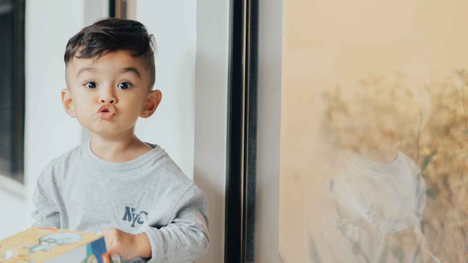 A child in a gray shirt stands indoors near a window, making a playful face while holding a colorful box.