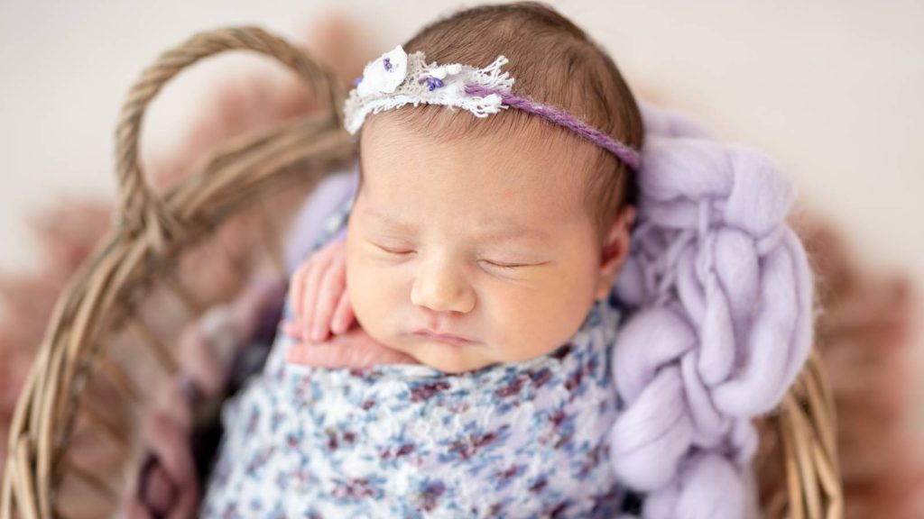 A sleeping baby wrapped in floral fabric is in a woven basket.