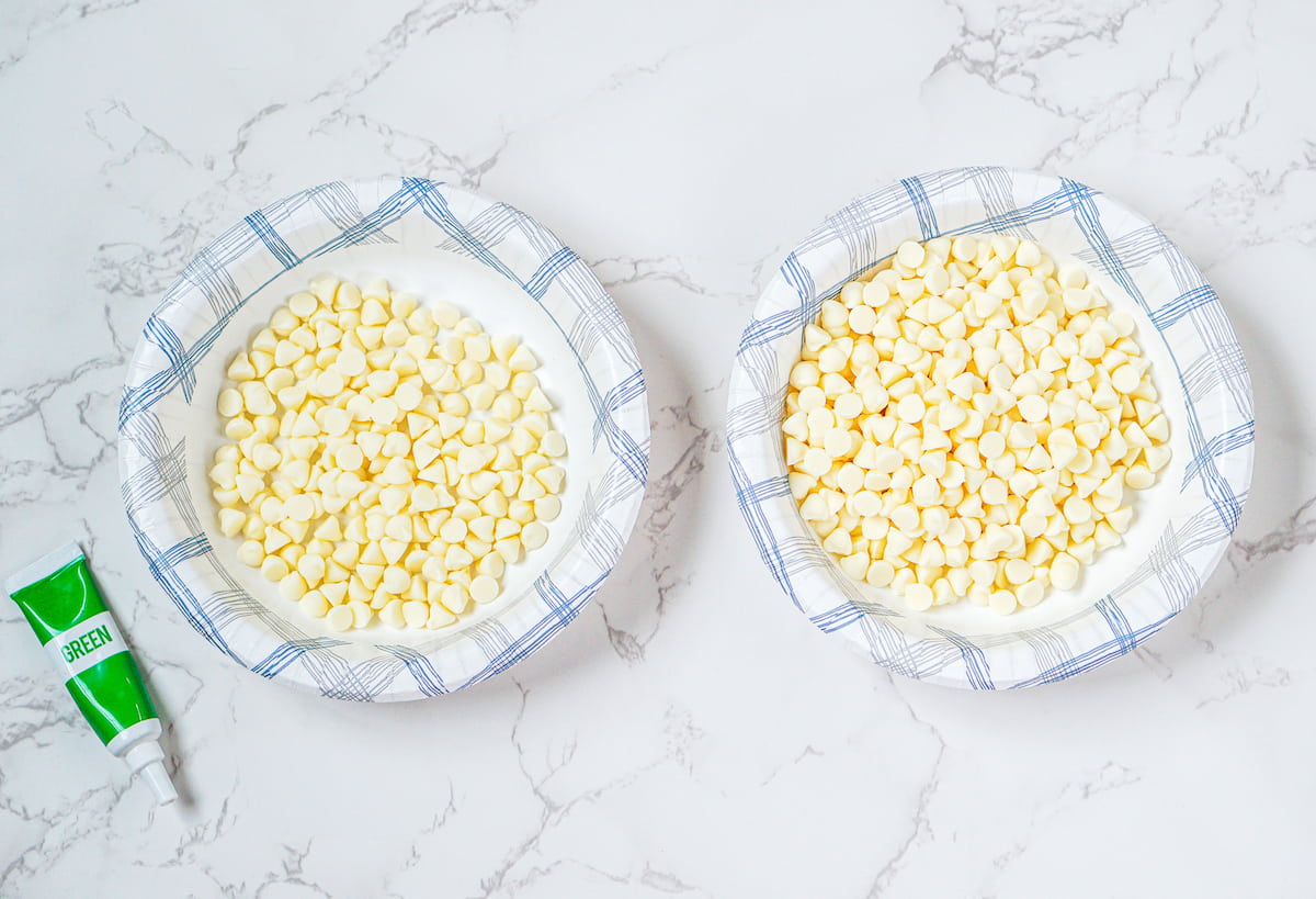 Two paper plates with white chocolate chips rest on a marble surface, ready to create festive leprechaun bark. A green food coloring tube sits nearby.