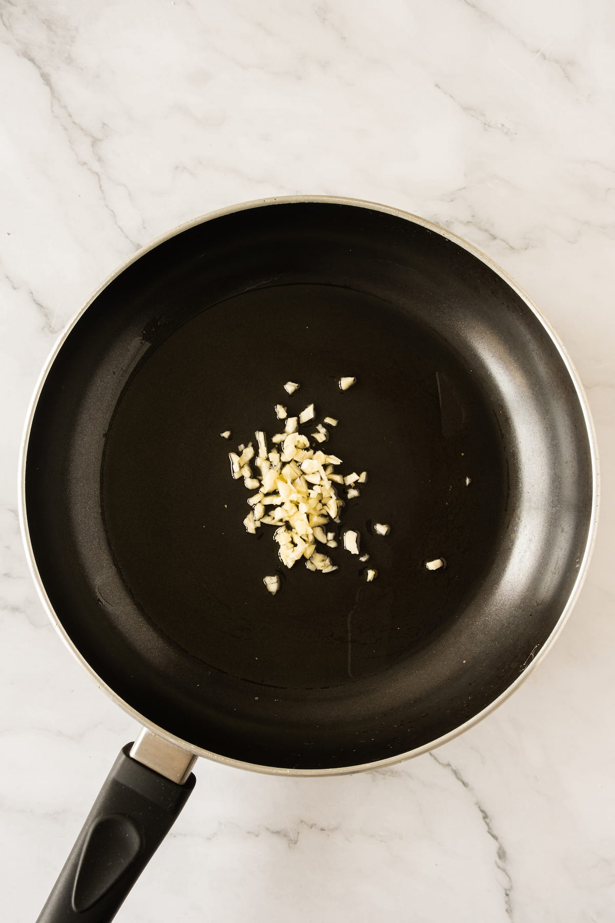 A small amount of chopped garlic sizzles in a black frying pan on a marble countertop.