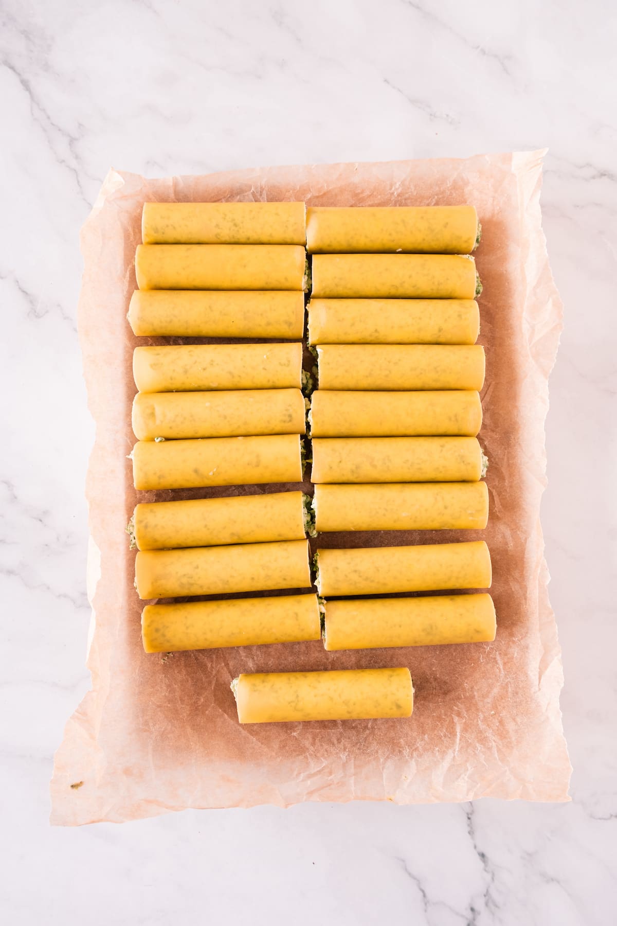 Uncooked cannelloni pasta tubes, stuffed with a vibrant spinach filling, are neatly arranged on parchment paper.
