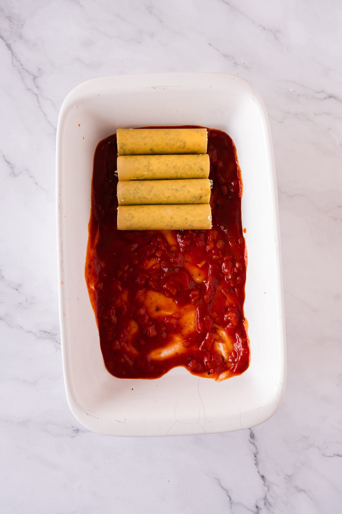 Four uncooked cannelloni, brimming with spinach and ricotta, rest on a thin layer of tomato sauce in a white baking dish atop a marble surface.