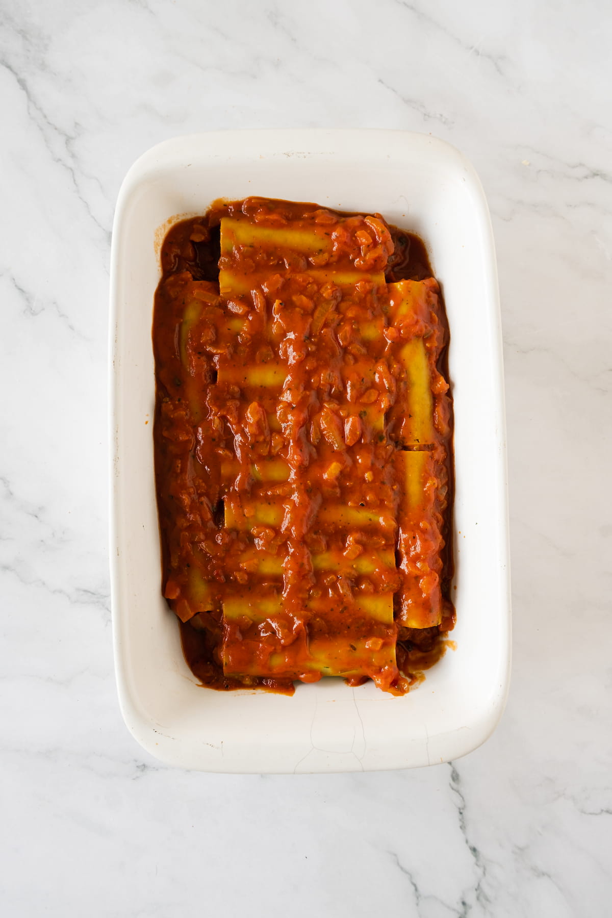 A marble surface cradles a baking dish filled with baked spinach cannelloni, smothered in a chunky red sauce.