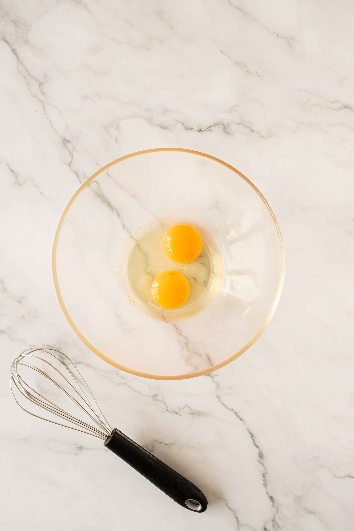 A glass bowl with two raw egg yolks and whites rests on a marble surface next to a metal whisk with a black handle.
