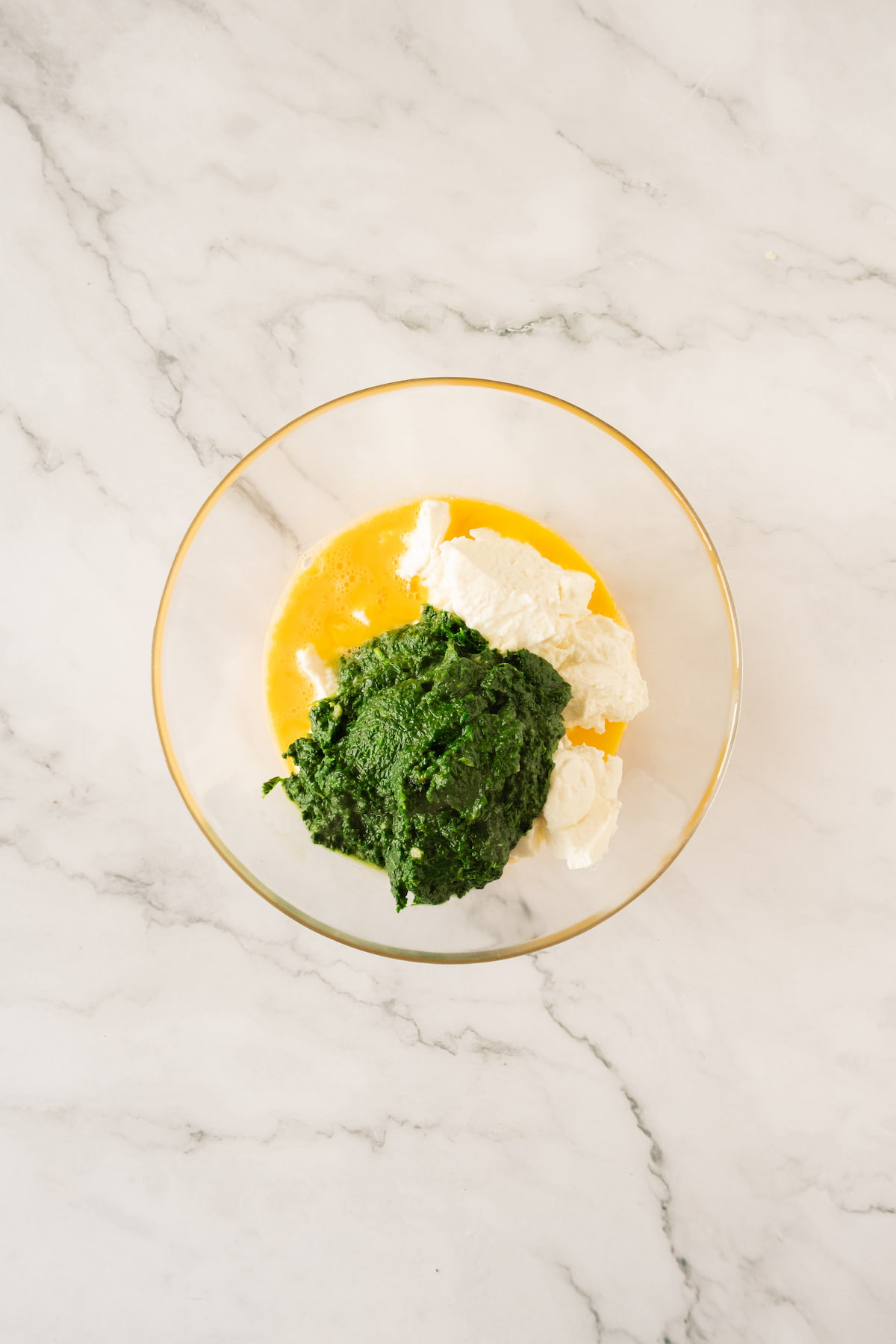 A glass bowl containing beaten eggs, ricotta, and spinach sits on a marble surface.