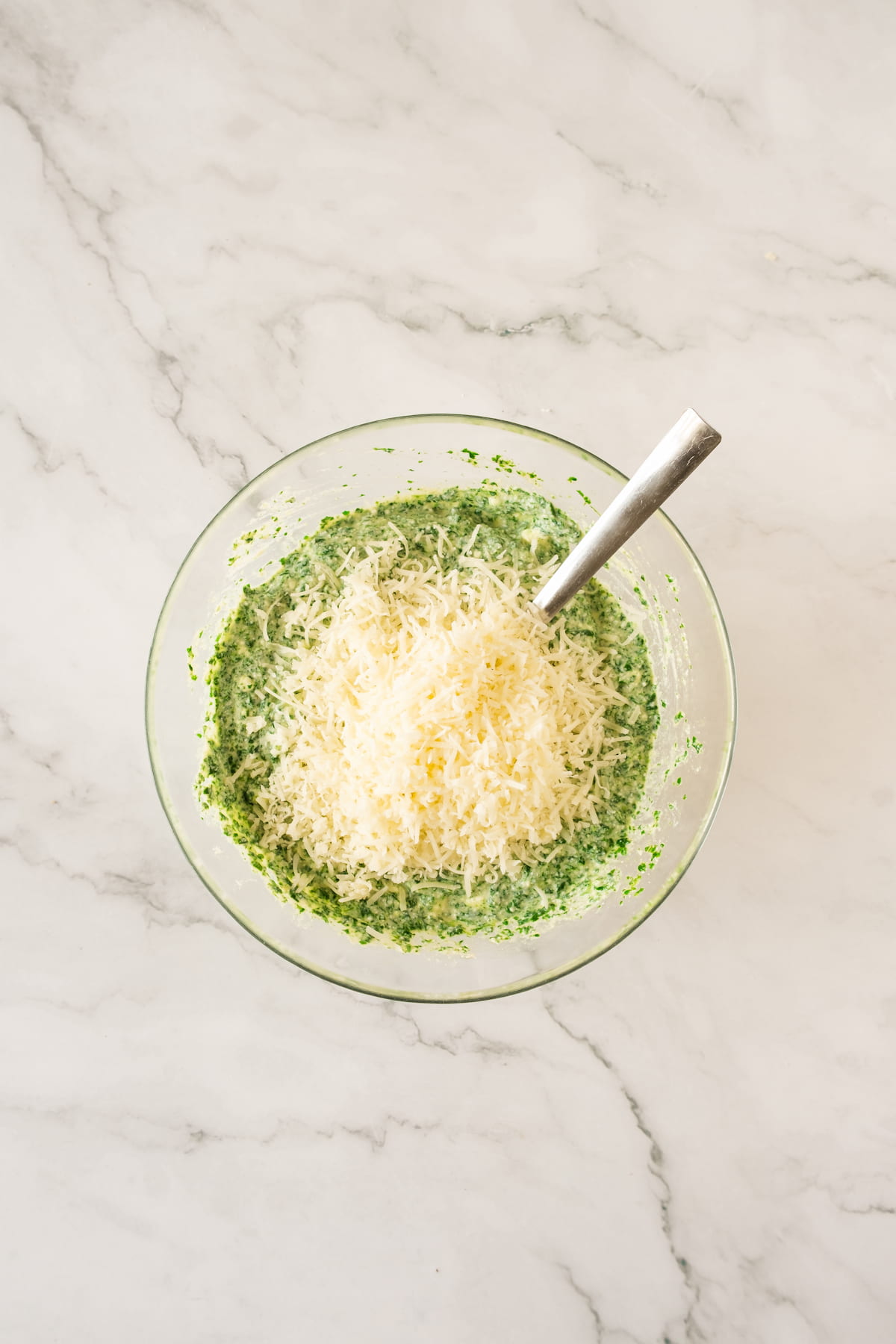 A bowl of creamy green spinach mixture is topped with grated Parmesan cheese. It sits invitingly on a marbled surface with a spoon nestled inside.