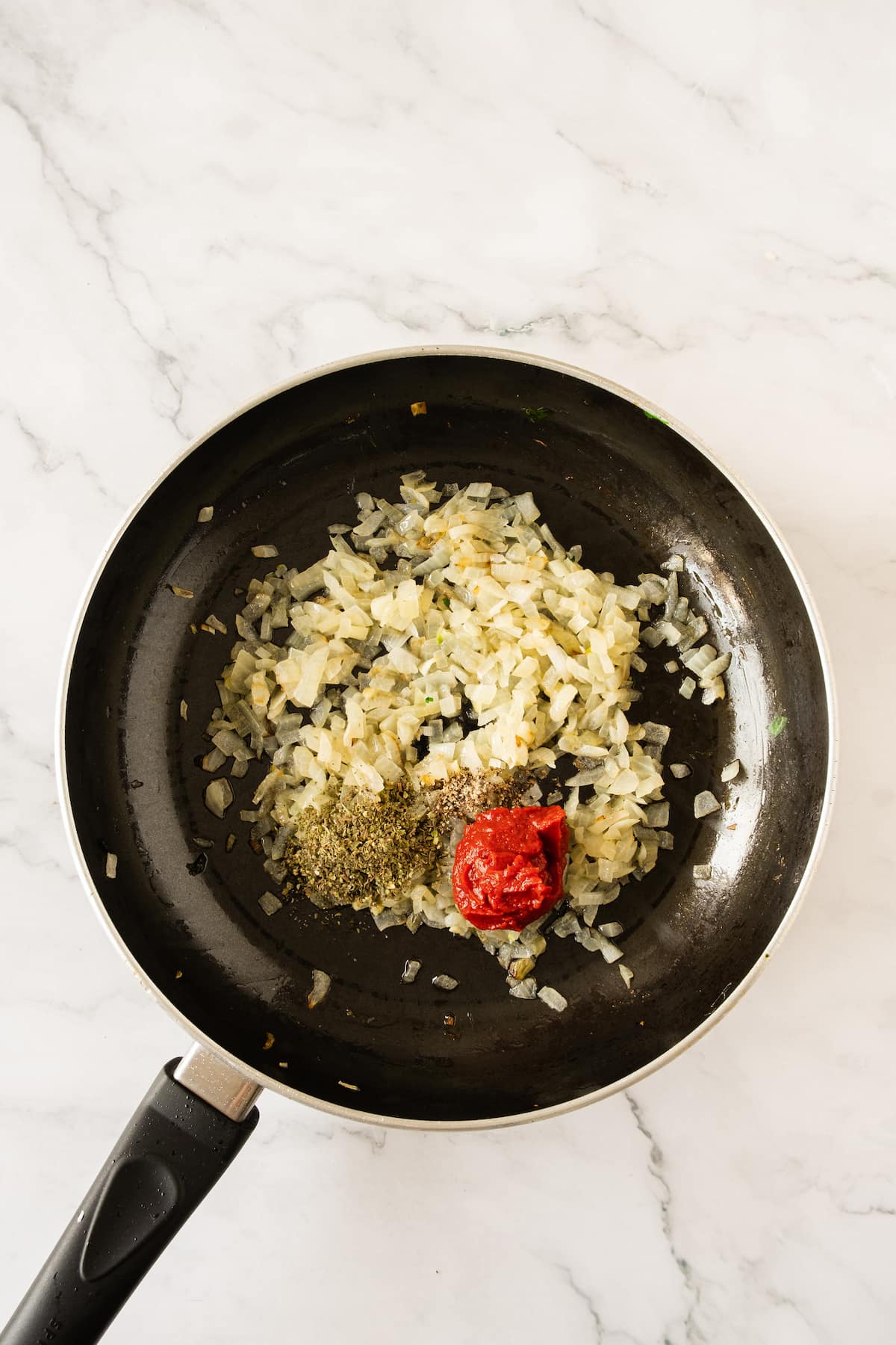 A frying pan with sautéed onions, tomato paste, and herbs on a marble surface, ready to complement a delicious baked spinach cannelloni.