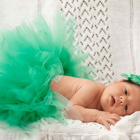 Baby lying on a white knitted blanket, wearing a green tulle skirt and a matching headband with a green flower.