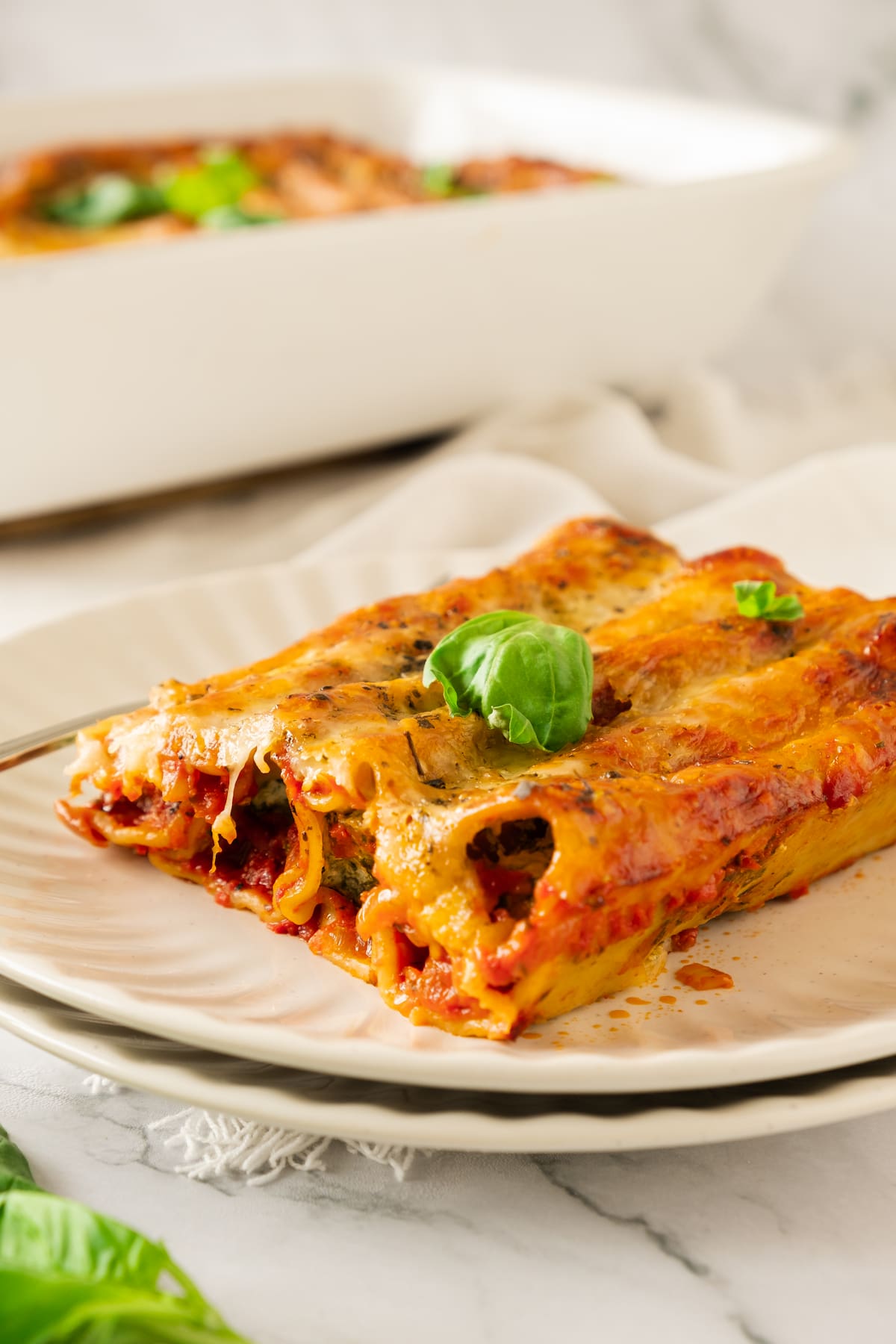 A plate of baked spinach cannelloni filled with creamy cheese, topped with melted cheese and a fresh basil leaf. In the background, a baking dish holds more delicious cannelloni.