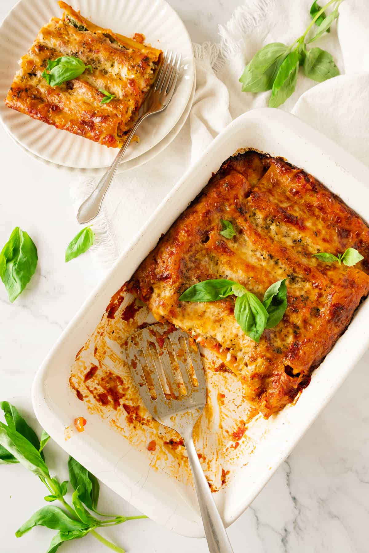 A rectangular dish of baked spinach cannelloni with slices removed, topped with basil leaves. A portion rests on a separate plate beside it, featuring an additional basil garnish for flavor contrast.