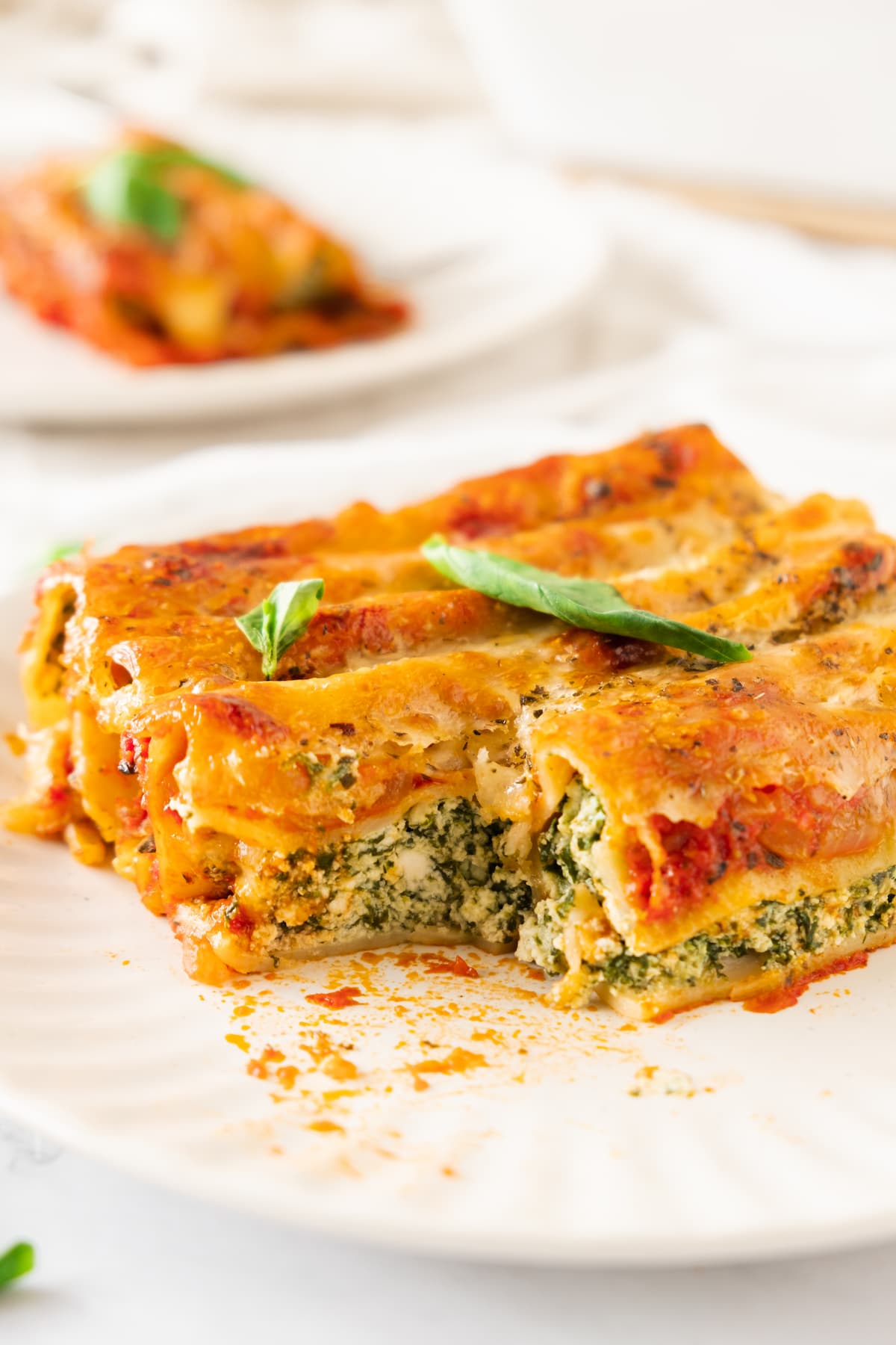 A slice of baked spinach cannelloni topped with melted cheese and garnished with basil sits elegantly on a white plate.