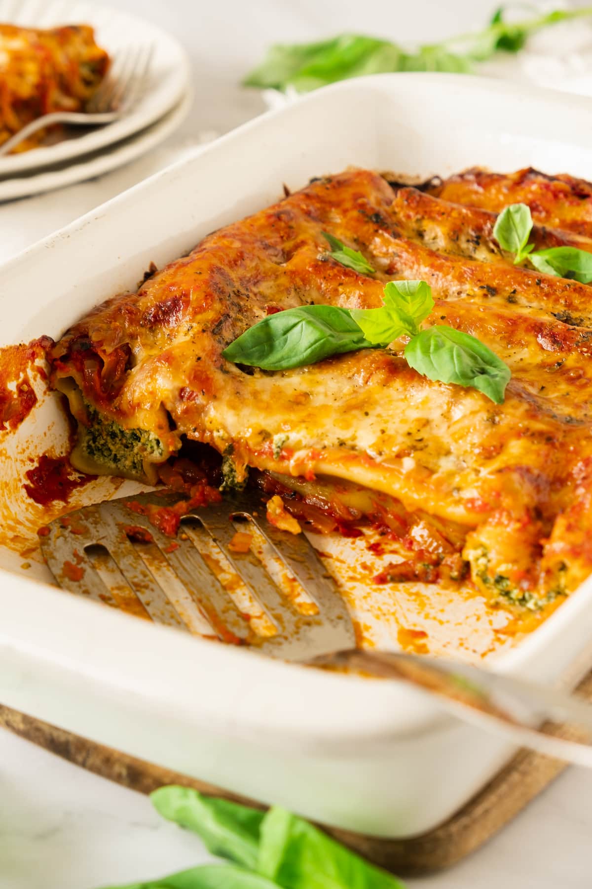 Baked spinach cannelloni in a white dish, topped with melted cheese and basil leaves. A portion is missing, with a serving spatula beside it. Nearby, plates await the deliciousness of baked spinach cannelloni for a perfect Italian feast.
