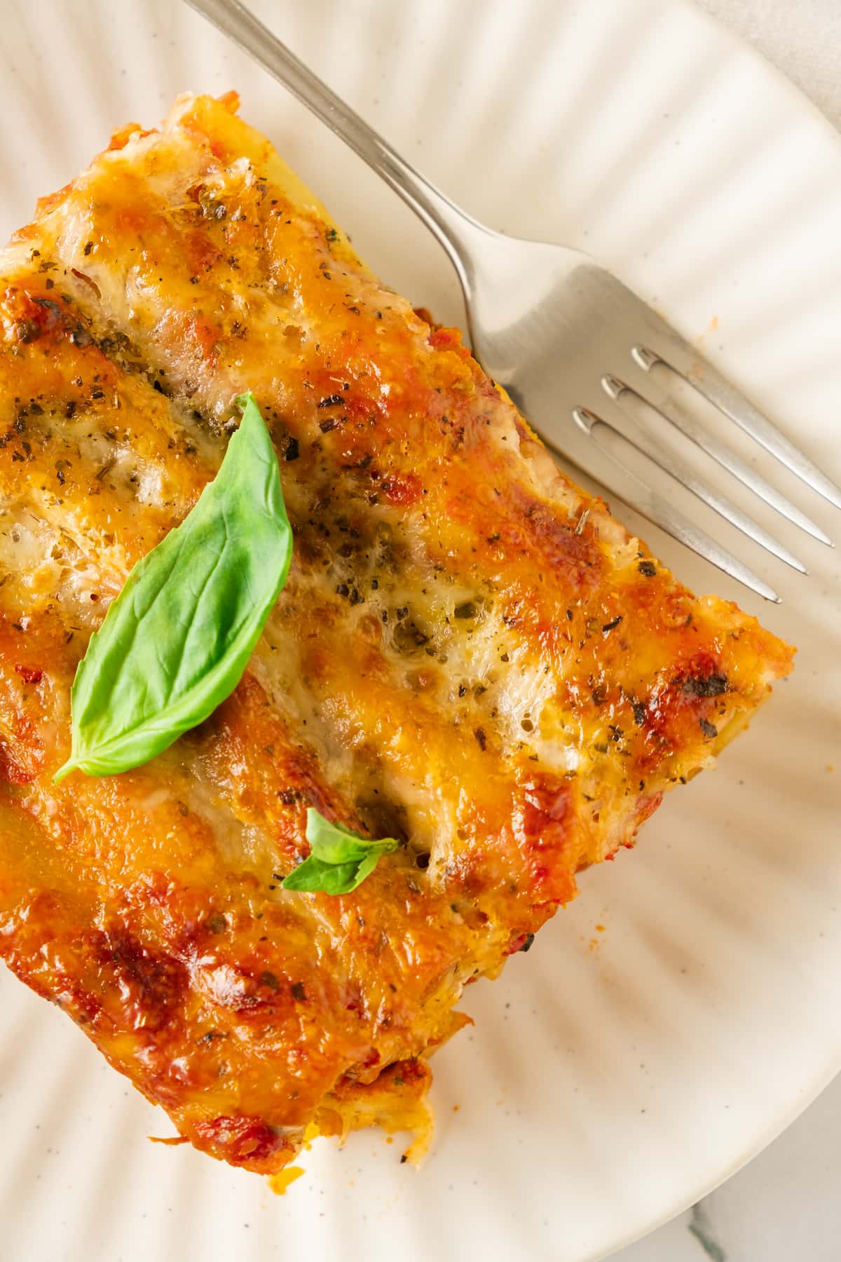 A slice of baked spinach cannelloni topped with basil sits on a white plate beside a fork.
