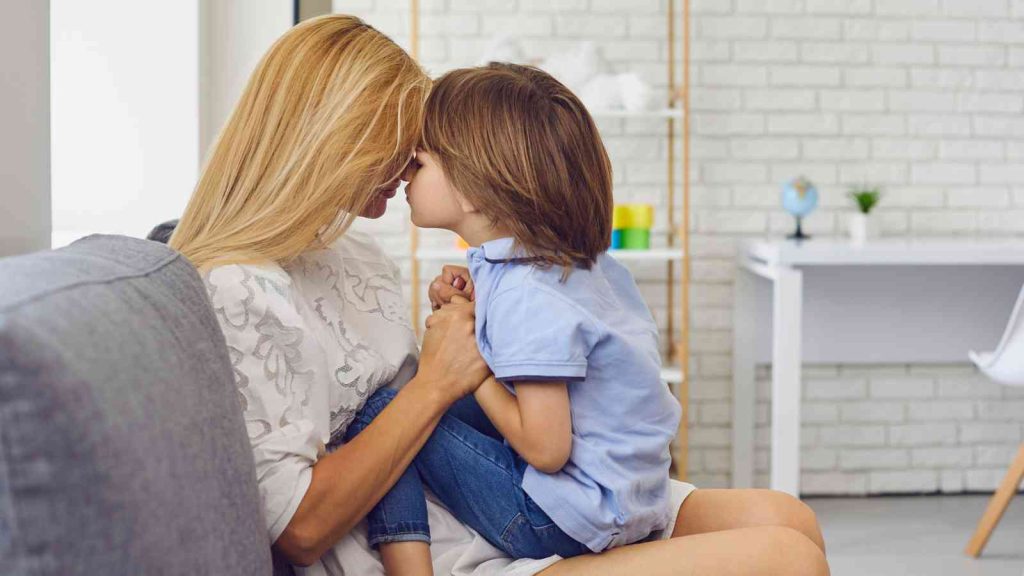 A woman and a child share a forehead touch while sitting on a couch in a bright room.
