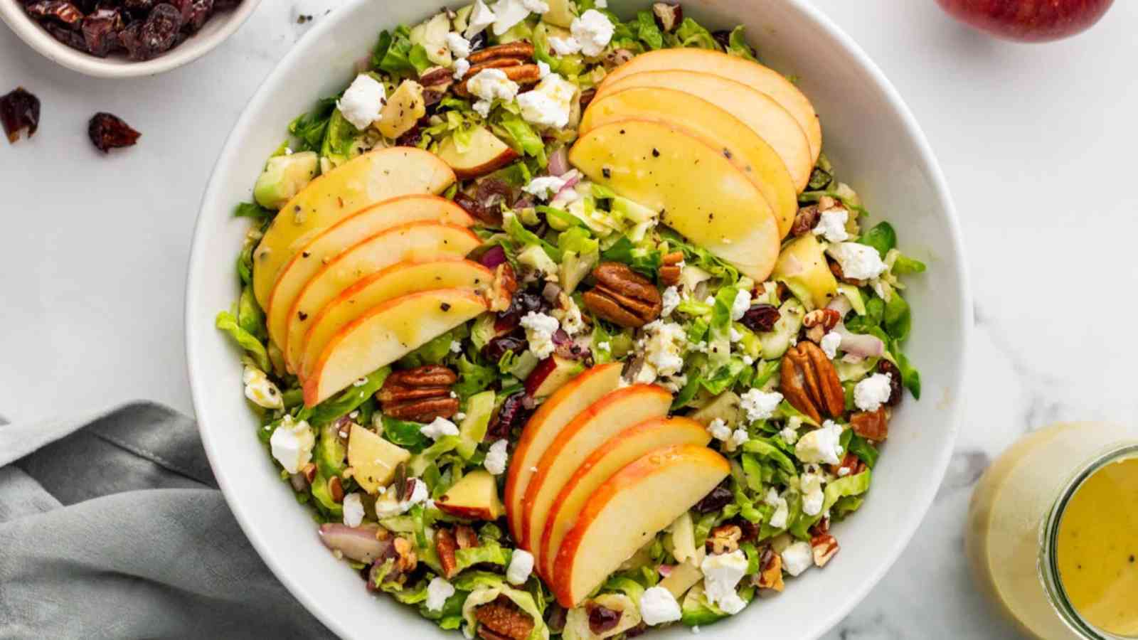 A salad with sliced apples, pecans, goat cheese, and leafy greens in a white bowl, with a side of vinaigrette dressing on a marble surface.