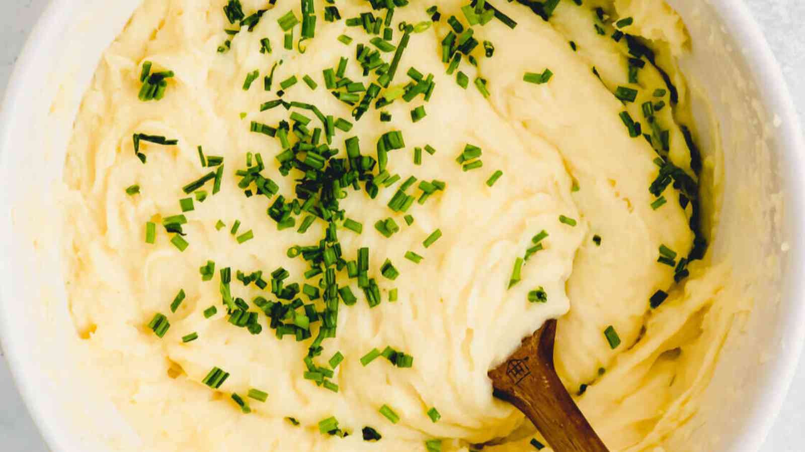 A bowl of mashed potatoes topped with chopped chives, with a wooden spoon resting in the mixture.