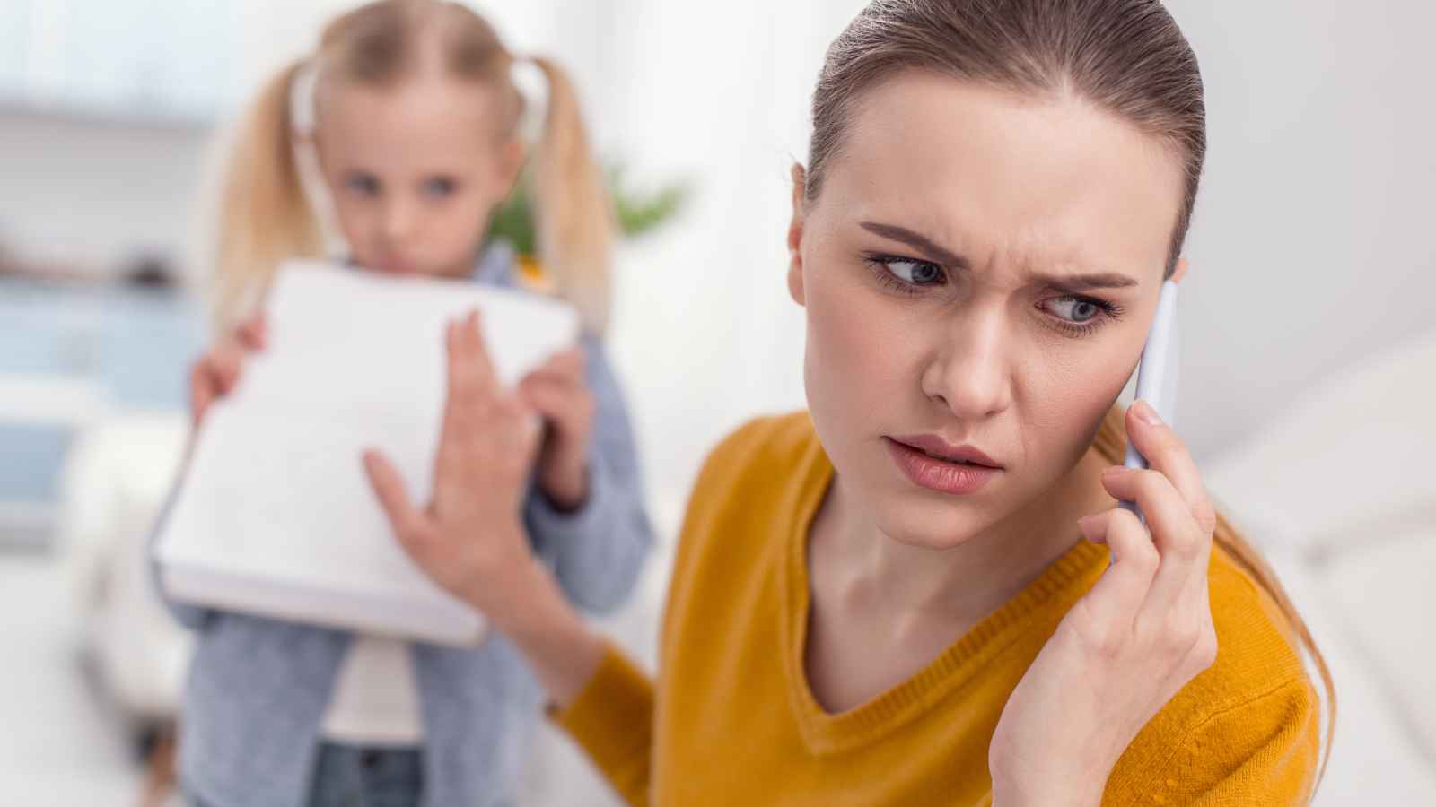 Woman on phone looking concerned, while a girl in the background holds an open book, appearing slightly blurred.
