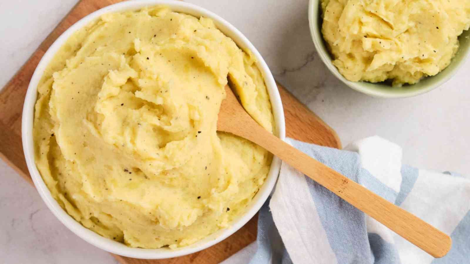 A bowl of mashed potatoes with a wooden spoon on a wooden board. A smaller bowl with more mashed potatoes is beside a striped cloth.