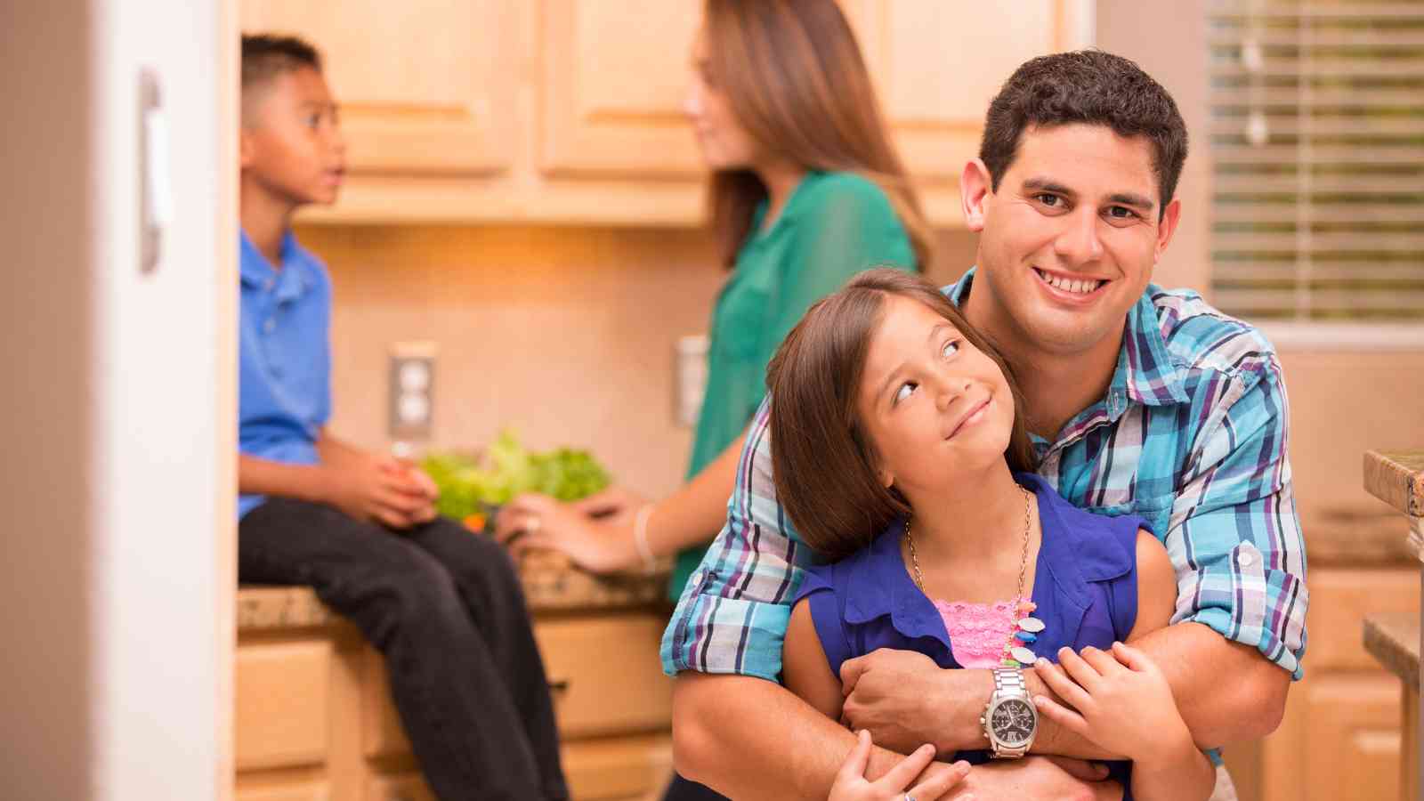 A man hugs a smiling girl in the foreground of a kitchen, while a woman and a boy sit in the background near the counter.