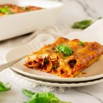 A slice of baked spinach cannelloni on a plate with a fork, garnished with basil, sits invitingly beside a baking dish brimming with deliciousness. In the background, baked spinach cannelloni adds its own charm to the scene, promising an irresistible Italian feast.