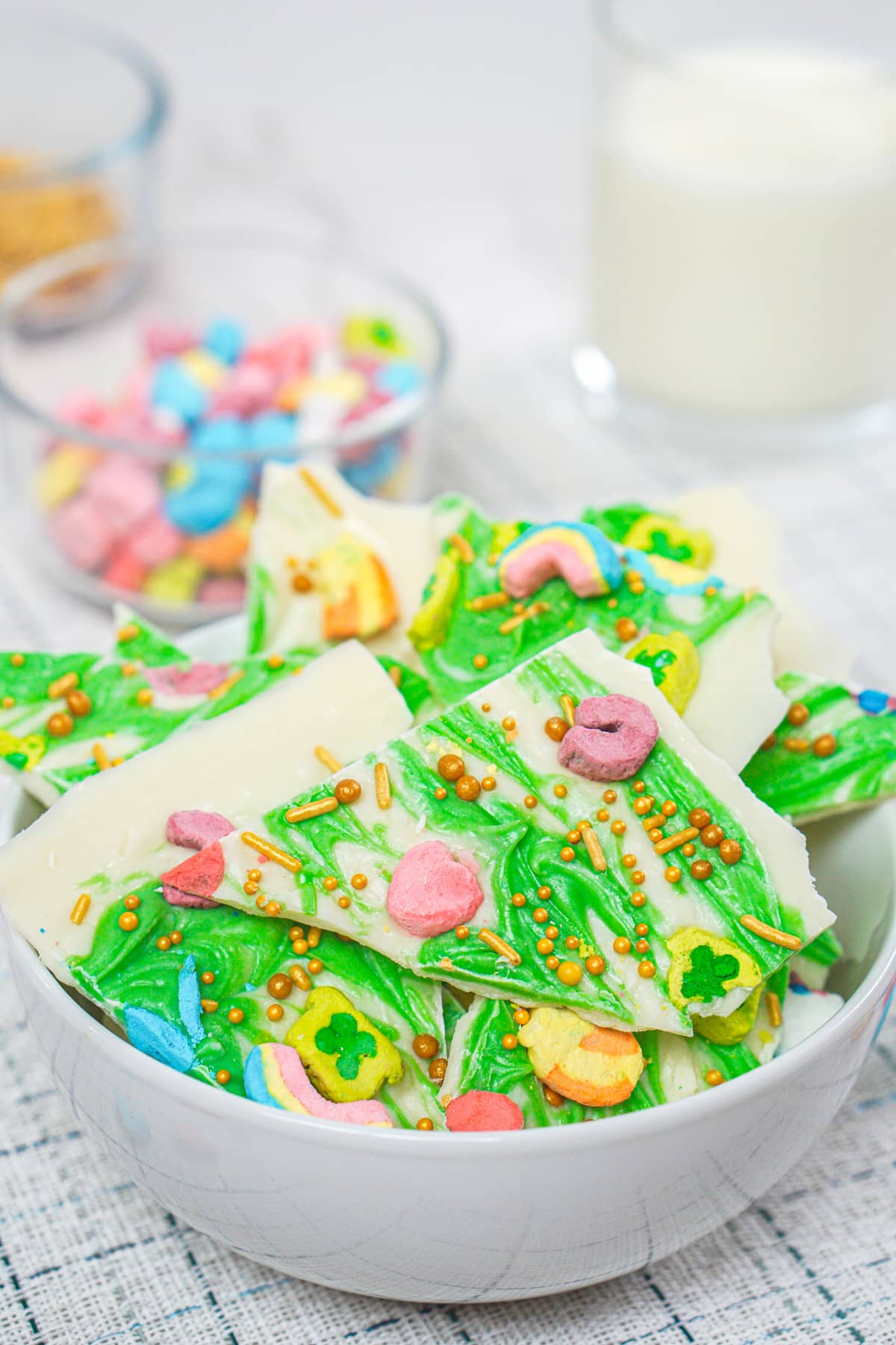 A bowl of leprechaun bark, crafted with green and white chocolate, is adorned with vibrant marshmallow pieces and shimmering gold sprinkles.